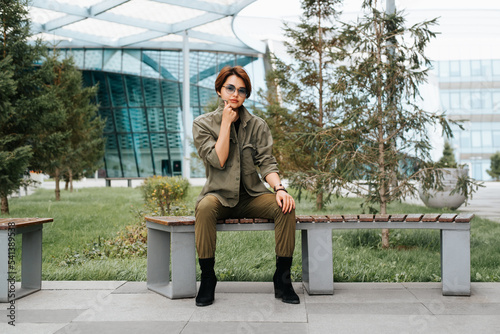 Asian young woman model with short hairstyle wearing glasses and casual green clothes posing and looking at camera while sitting on a bench in a modern neighborhood