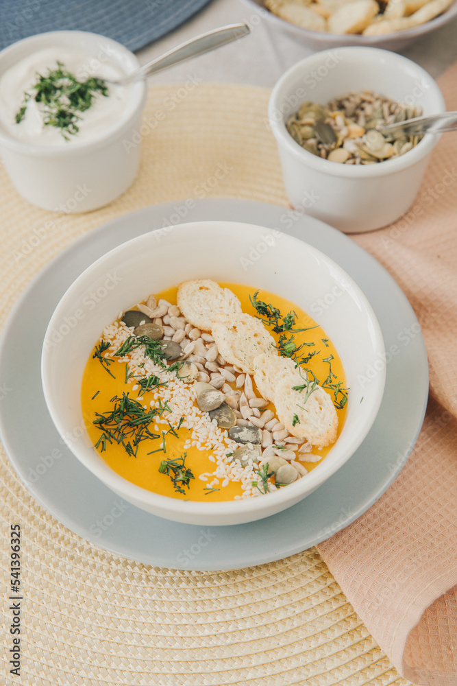 A white bowl with pumpkin cream soup garnished with pumpkin, sesame and sunflower seeds, crackers and dill. In the background, in blur, pumpkin seeds in bowl and sour cream in bowl. A white bowl with pumpkin cream soup garnished with pumpkin, sesame and sunflower seeds, crackers and dill. In the background, in blur, pumpkin seeds in bowl and sour cream in bowl.