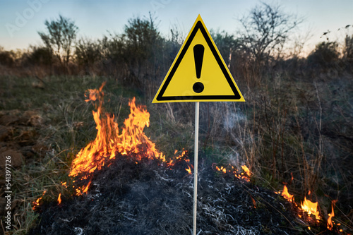 Burning dry grass and warning sign with exclamation mark warns about dangerous in field with fire. Concept of ecology and natural disaster.