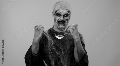 Zombie man with make-up with fake wounds scars celebrate success win scream rejoices doing winner hands gesture say Yes in gray studio room. Sinister undead guy. Halloween, filming, staging concept