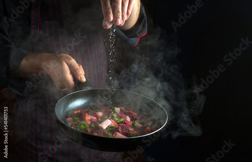 Fototapeta Naklejka Na Ścianę i Meble -  The chef salts vegetables in a hot frying pan. Menu concept for hotel or restaurant on black background