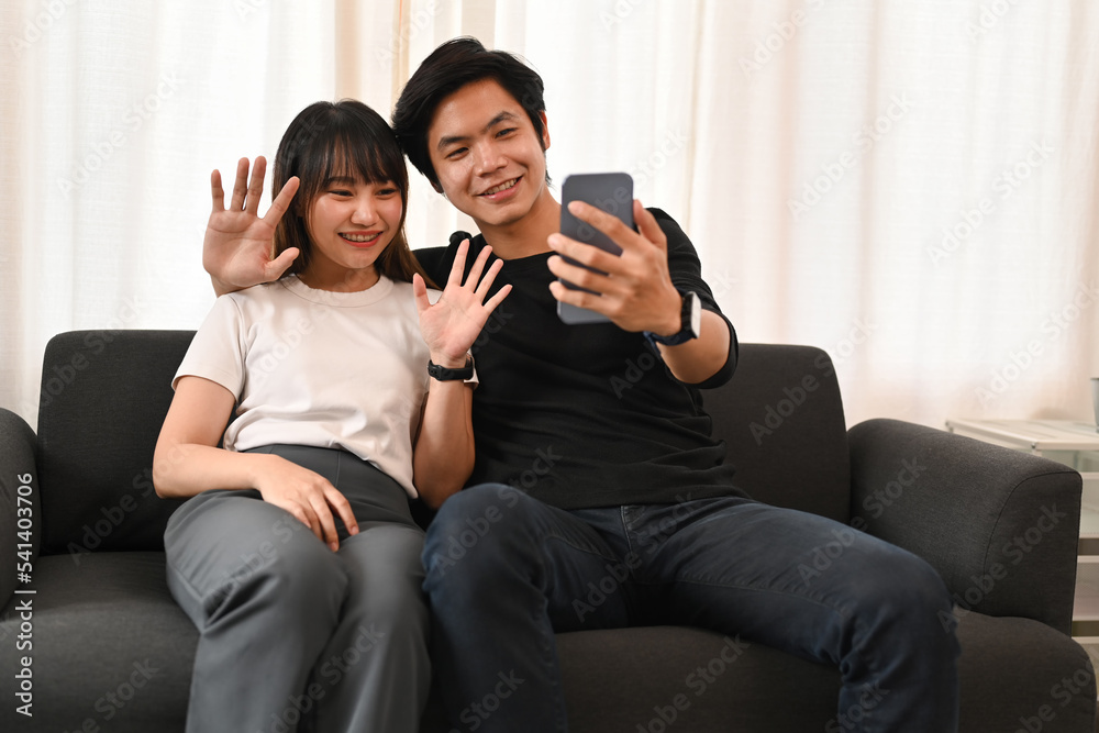 Fototapeta premium Happy young couple taking selfie, making video call on smart phone while sitting on couch at home