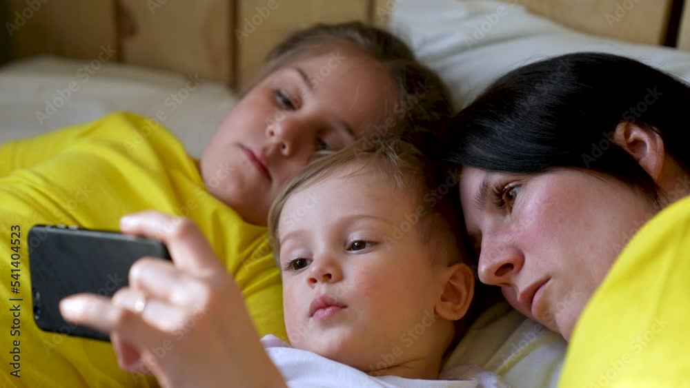 Happy family concept. Mom, daughter and son are play on bed. Child watching video on smartphone. Family leisure at home. Boy and girl with mom are watching movie on smartphone. Home education concept