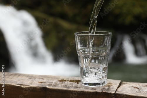 Fresh water pouring into glass on wooden surface near waterfall. Space for text © New Africa