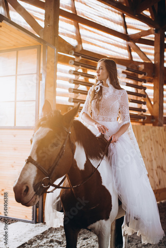 A charming boho bride rides a horse on a ranch at sunset in winter.