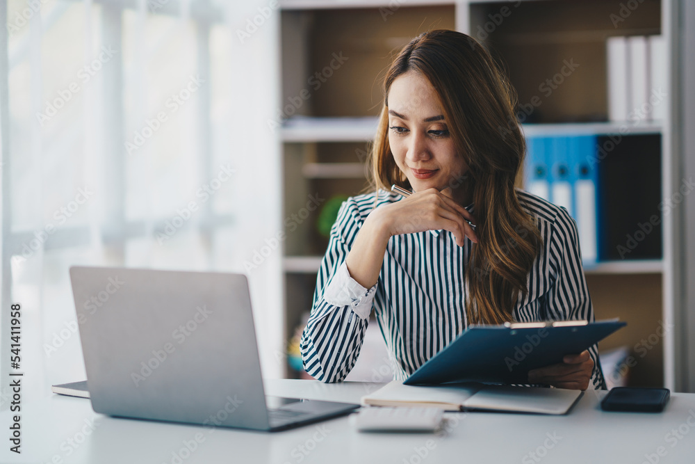 Φωτογραφία Portrait of a cheerful Asian businesswoman using a laptop and writing on a charg
