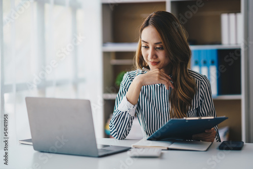 Quadro su tela Portrait of a cheerful Asian businesswoman using a laptop and writing on a charg