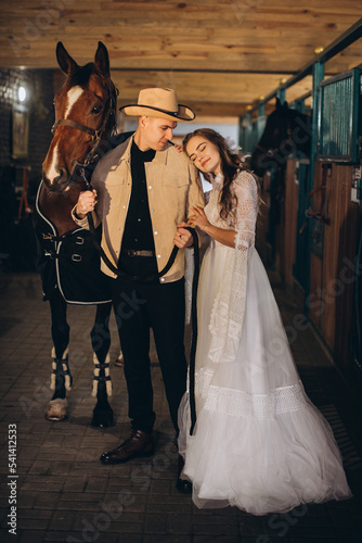 Handsome young cowboy man and his charming boho bride pose in a stable with horses.