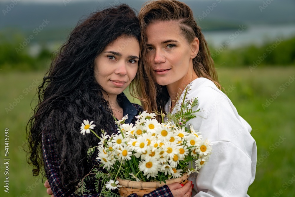 Two women enjoy nature in a field of daisies. Girlfriends hugging hold a bouquet of daisies and look at the camera. Two women enjoy nature in a field of daisies. Girlfriends hugging hold a bouquet of daisies and look at the camera.