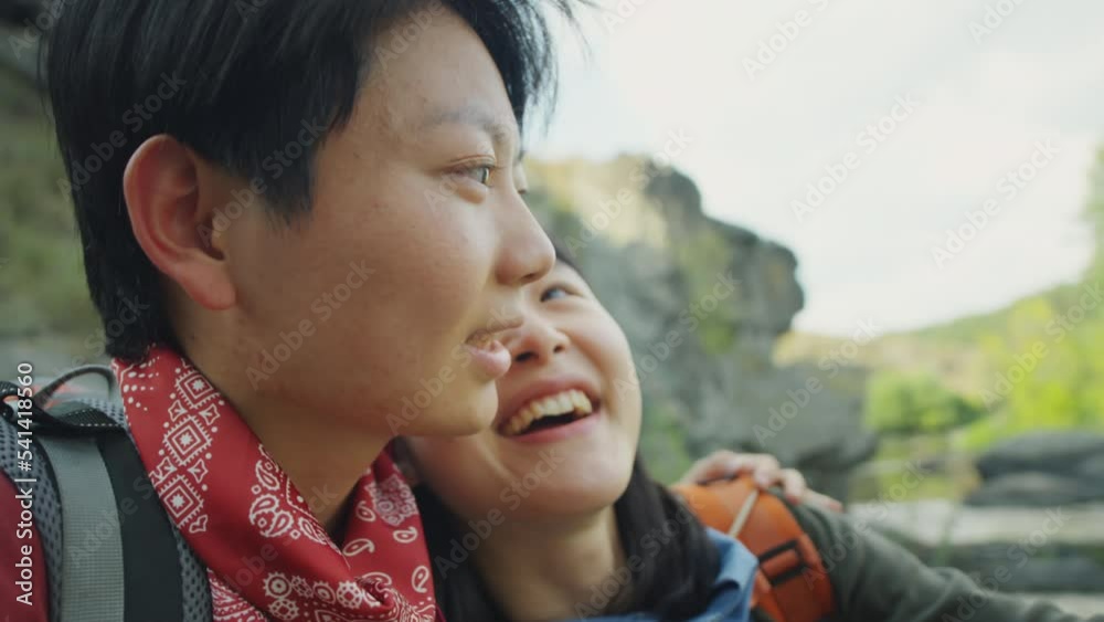 Close up shot of happy Asian female couple embracing, smiling and chatting while resting on river embankment during summer hike Close up shot of happy Asian female couple embracing, smiling and chatting while resting on river embankment during summer hike