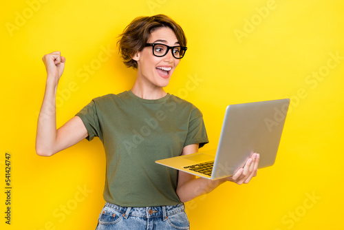 Photo of impressed positive woman bob hairdo dressed khaki t-shirt hold laptop clench fist shout yes isolated on yellow color background