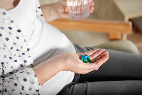Pregnant woman holding pile of pills and glass with water indoors, closeup