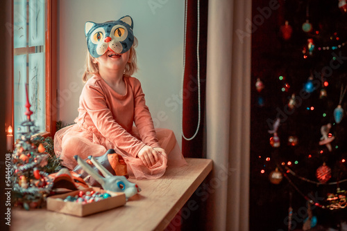 Cute little girl is dressing up a tiny Christmas tree and trying on New Year's animal masks. Image with selective focus and toning. 
