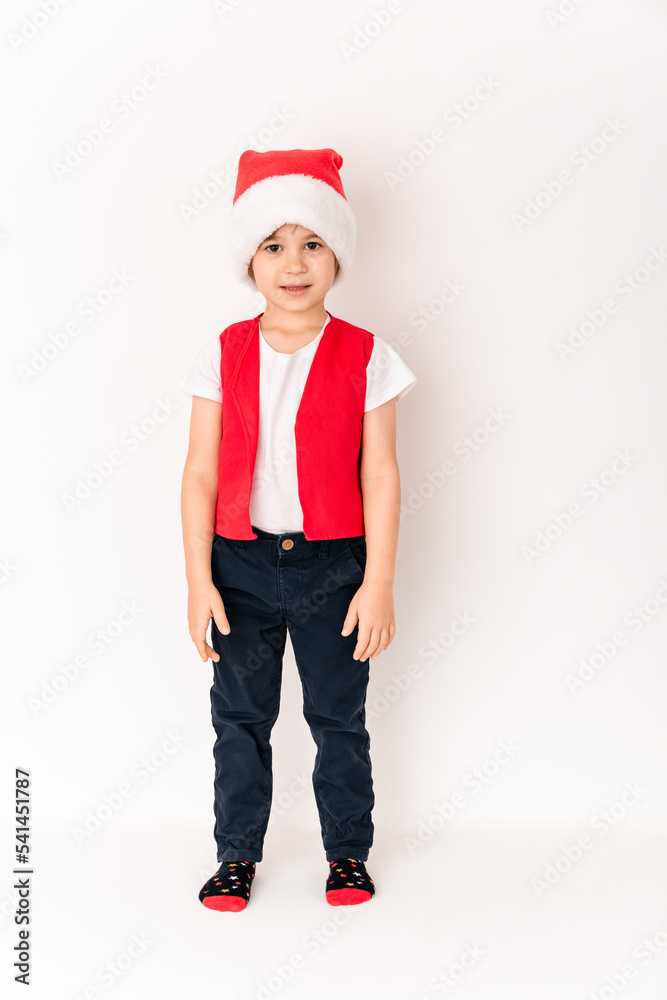 Portrait of standing smiling child in red Santa Claus hat looking at camera isolated on white background. 5 year happy European boy. Greeting card mockup design. New Year holiday atmosphere concept Portrait of standing smiling child in red Santa Claus hat looking at camera isolated on white background. 5 year happy European boy. Greeting card mockup design. New Year holiday atmosphere concept