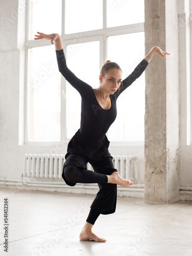 Fototapeta Naklejka Na Ścianę i Meble -  Modern female dancer in black outfit balancing on one leg while practices in a dance studio
