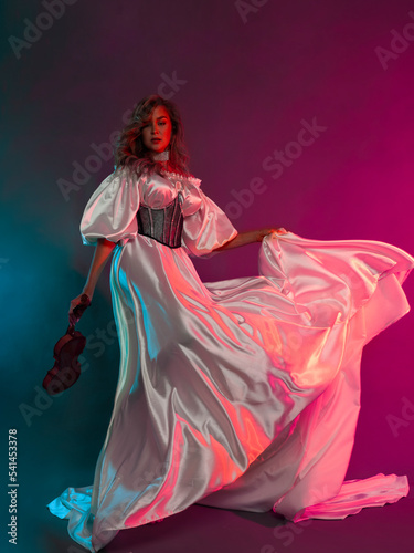 A woman poses in a long white silk dress on a colored background with a violin in her hands. A model in a flowing dress. Portrait of a full-length beauty.