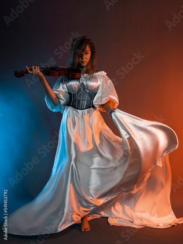 A woman poses in a long white silk dress on a colored background with a violin in her hands. A model in a flowing dress. Portrait of a full-length beauty.