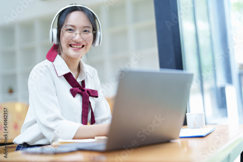 Portrait of a teenage Asian woman using a computer, wearing headphones and using a notebook to study online via video conferencing on a wooden desk in library