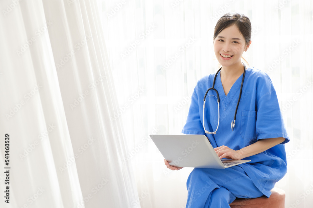 Portrait of a young Asian happy nurse wearing medical scrubs looking at the camera with a stethoscope and using a laptop while sitting with a white curtain in the background. Image with copy space. Portrait of a young Asian happy nurse wearing medical scrubs looking at the camera with a stethoscope and using a laptop while sitting with a white curtain in the background. Image with copy space.