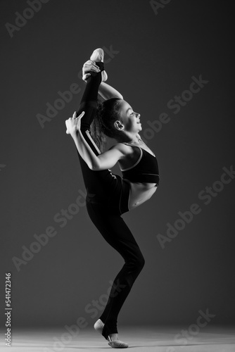 A young flexible girl gymnast in a tracksuit does gymnastic stretching exercises. She threw her leg over her head. Black and white photo on a white background.