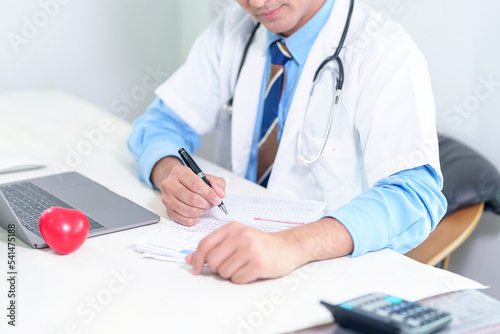 Senior doctor physician using laptop, writing prescription with stethoscope on desk. Old male professional therapist wearing white coat working in hospital. Healthcare, medicine concept. Close up view