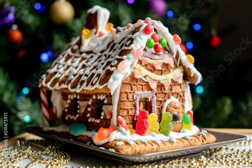 Homemade gingerbread house decorated with icing, sweets , and jelly candies with blurred Christmas tree on background.