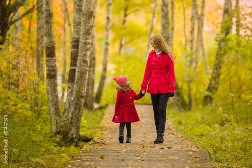 Happy little daughter and young adult mother walking on trail at birch tree forest. Spending time together in beautiful colorful autumn day. Enjoying stroll. Front view.