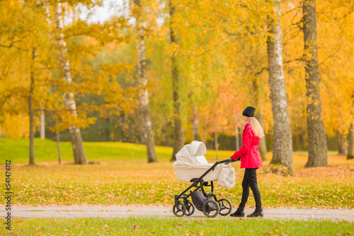 Young adult mother in red coat pushing white baby stroller and walking at town park in colorful autumn day. Spending time with newborn and breathing fresh air. Enjoying stroll. Side view.