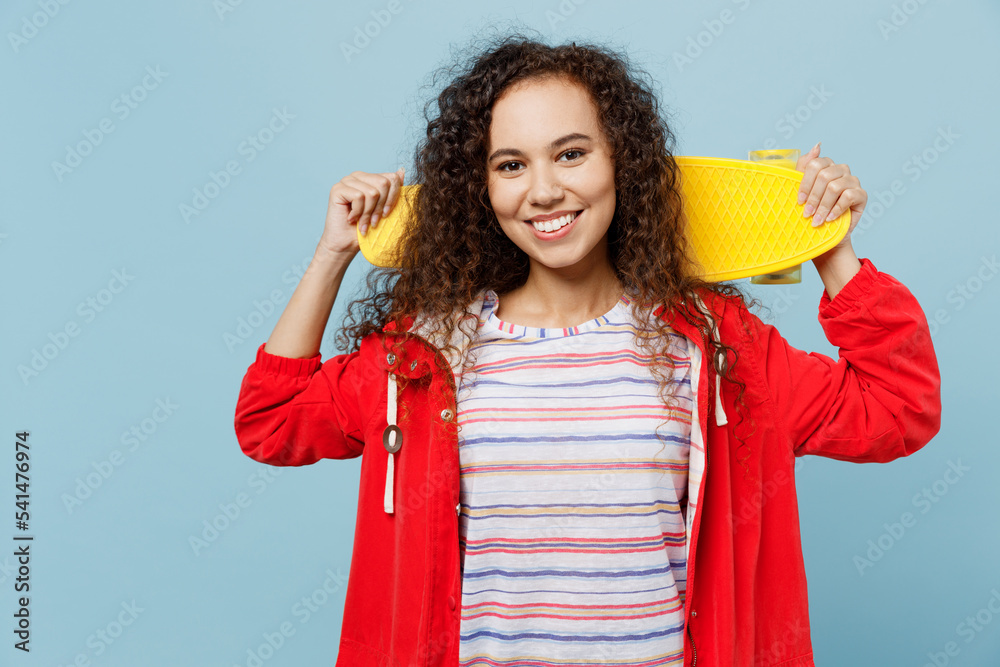 Young smiling woman of African American ethnicity 20s she wear red jacket hold skateboard pennyboard behind neck isolated on plain pastel light blue cyan background. Wet fall weather season concept. Young smiling woman of African American ethnicity 20s she wear red jacket hold skateboard pennyboard behind neck isolated on plain pastel light blue cyan background. Wet fall weather season concept.
