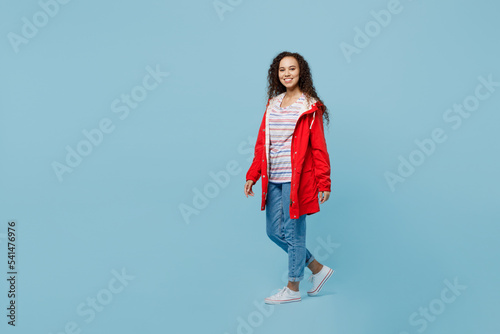 Full body sideways happy fun young woman of African American ethnicity 20s she wear red jacket walk go look camera isolated on plain pastel light blue cyan background. Wet fall weather season concept.
