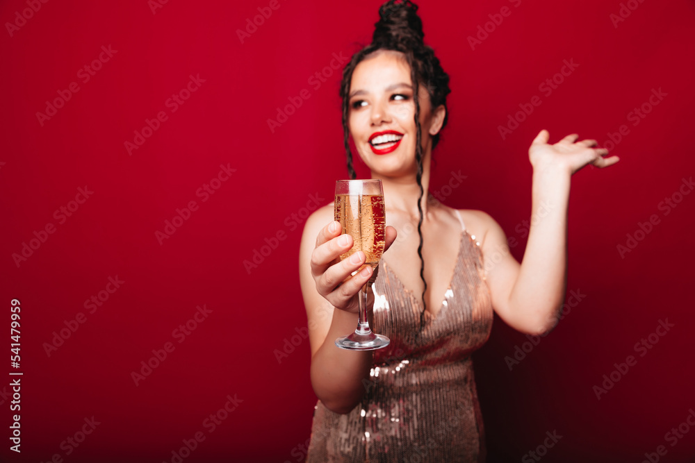 A smiling woman in an evening dress enjoys a party, holding a bottle of champagne and raising a glass on a red background.a woman celebrates her birthday A smiling woman in an evening dress enjoys a party, holding a bottle of champagne and raising a glass on a red background.a woman celebrates her birthday
