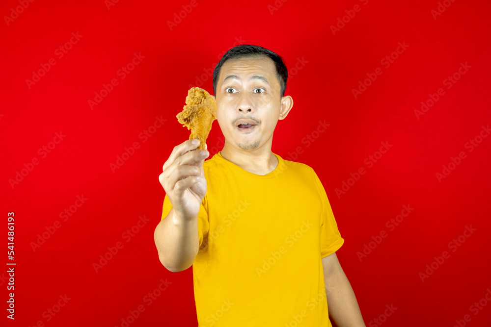 Portrait of young handsome Asian man looking at fried chicken in his hands, wearing yellow shirt and red background. Portrait of young handsome Asian man looking at fried chicken in his hands, wearing yellow shirt and red background.