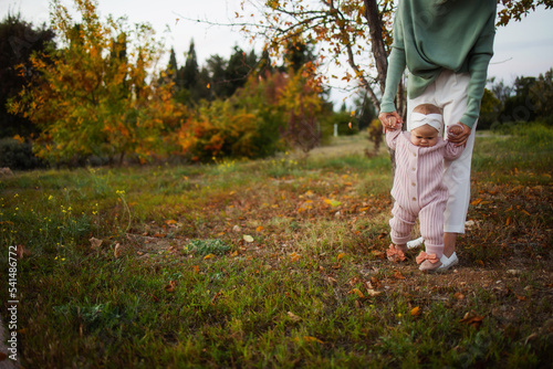Young mother walks and holds her child's hands in an autumn deciduous park. Stylish mother teaches her child to walk. Taking care of my daughter. Daughters Day © Елена Бабанова