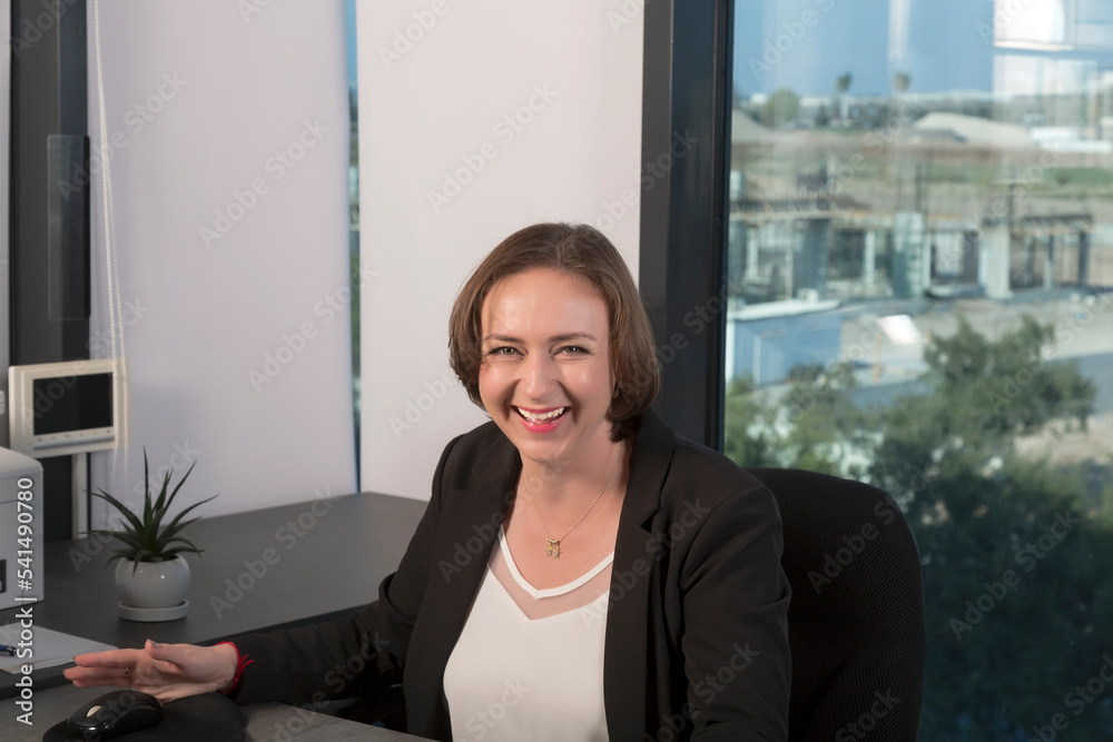 Caucasian smiling brunette model at work in the office and looking at camera. Beautiful young adult business woman with loose brown hair in black suit with professional make up. working behind desk Caucasian smiling brunette model at work in the office and looking at camera. Beautiful young adult business woman with loose brown hair in black suit with professional make up. working behind desk