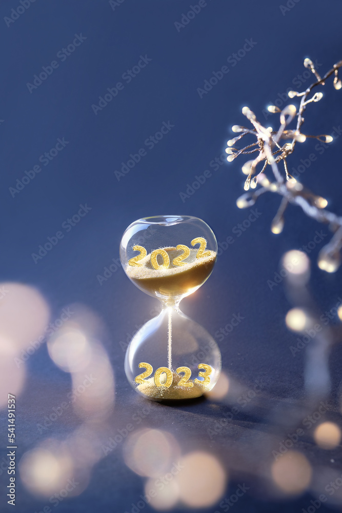 End of the year 2022, Silverster, Happy New Year 2023. Hourglass with numbers 2022 and 2023 and light garland on dark blue background. Hourglass is also known as sandglass, sand glass, sand timer or - obrazy, fototapety, plakaty 