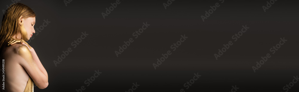 Fototapeta premium Side view of fair haired model in golden paint standing isolated on grey, banner