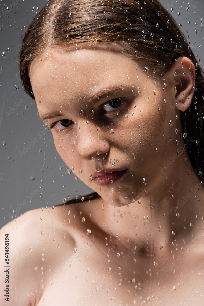 Fair haired model with naked shoulder looking at camera behind wet glass on grey background
