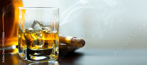 A bottle and a glass of whiskey with ice and a steaming Cuban cigar on a table against a light background. Men's club banner idea. Copy space for text