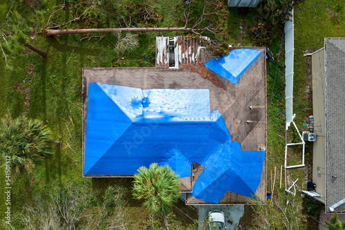 Aerial view of damaged in hurricane Ian house roof covered with blue protective tarp against rain water leaking until replacement of asphalt shingles