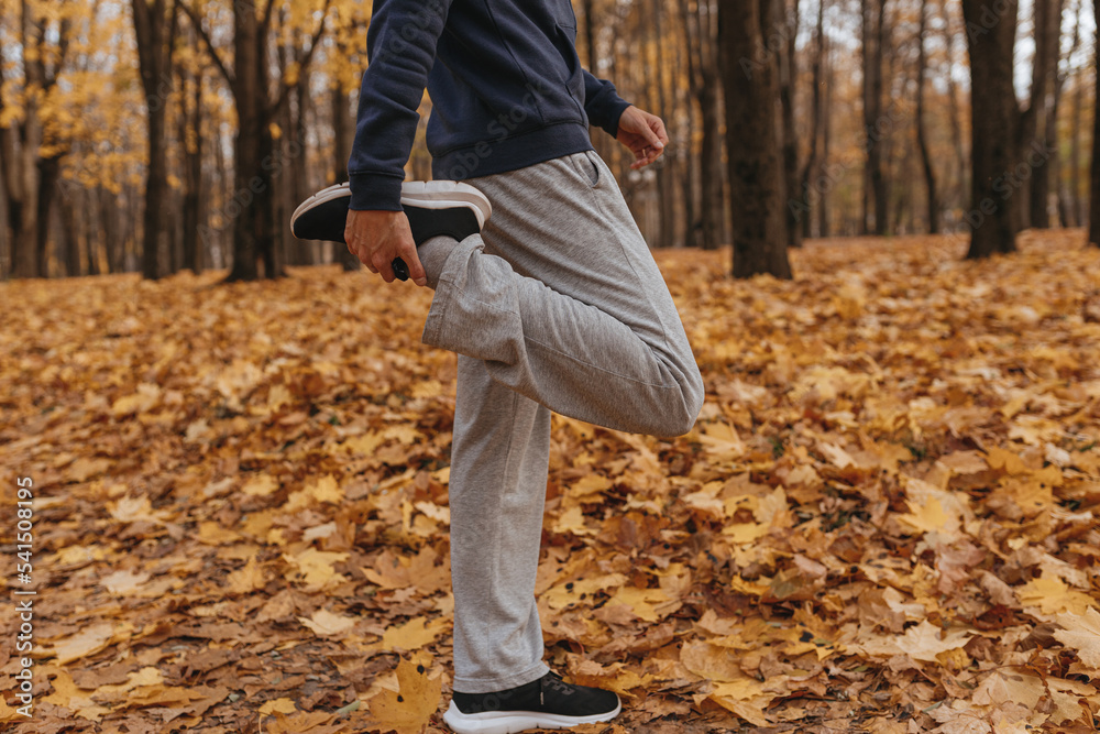 Side view of crop male athlete in sportswear stretching leg while standing in autumn park and doing exercise during outdoor fitness training Side view of crop male athlete in sportswear stretching leg while standing in autumn park and doing exercise during outdoor fitness training