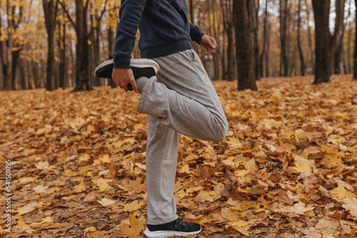 Side view of crop male athlete in sportswear stretching leg while standing in autumn park and doing exercise during outdoor fitness training 