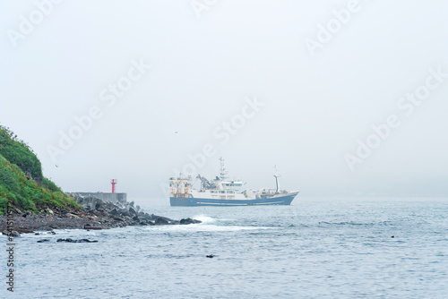 fishing vessel emerges from behind a cape with a lighthouse, sailing into a foggy sea