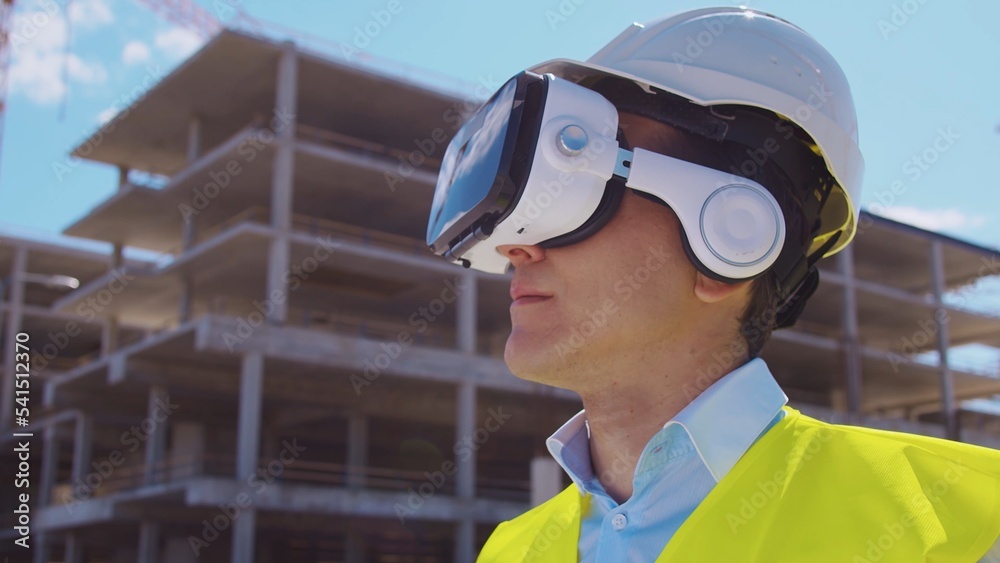 Professional builder in VR helmet standing in front of construction site and using virtual and augmented reality technologies. Office building and crane background. Real estate and investment.