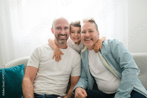 Happy two man couple with adopted child sit on sofa at home