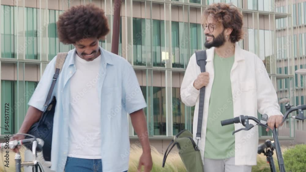 Two diverse young male smiling and talking while walking their bikes in ...