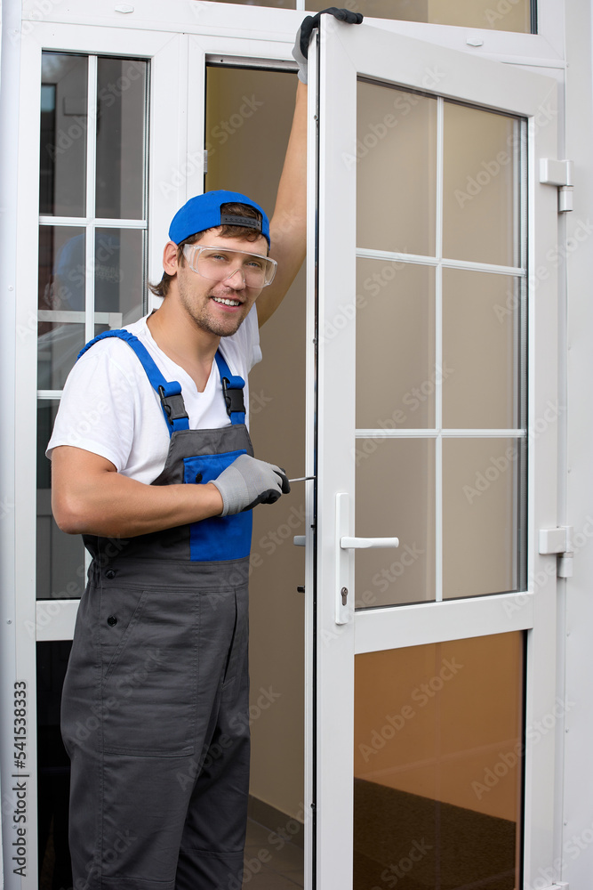 Tall male worker, smiling, stands at open door in overalls and protective glasses, repairing door lock with a screwdriver. An experienced master performs maintenance work on locks on a plastic door Tall male worker, smiling, stands at open door in overalls and protective glasses, repairing door lock with a screwdriver. An experienced master performs maintenance work on locks on a plastic door