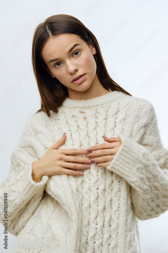 a beautiful, attractive playful woman in a white knitted sweater stands holding her fingers together on her chest on a bright studio background and looks merrily at the camera a beautiful, attractive playful woman in a white knitted sweater stands holding her fingers together on her chest on a bright studio background and looks merrily at the camera