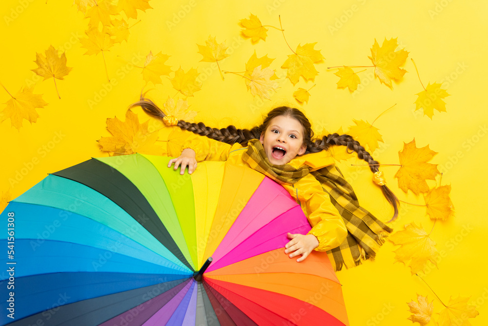A child holds onto an autumn multicolored umbrella lying on a yellow background with maple fallen leaves. A little girl in a raincoat with a scarf is enjoying the autumn weather. A child holds onto an autumn multicolored umbrella lying on a yellow background with maple fallen leaves. A little girl in a raincoat with a scarf is enjoying the autumn weather.