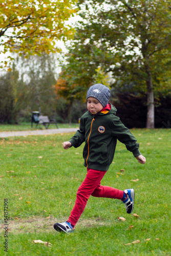 A boy wearing a green jacket and a cap running on a lawn in a park on an autumn evening