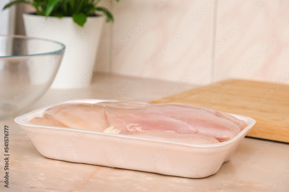 Defrosted raw chicken fillet in a disposable white tray. Raw chicken breast for cooking chopped chicken cutlets. Glass bowl and wooden cutting board for slicing chicken fillet. Defrosted raw chicken fillet in a disposable white tray. Raw chicken breast for cooking chopped chicken cutlets. Glass bowl and wooden cutting board for slicing chicken fillet.